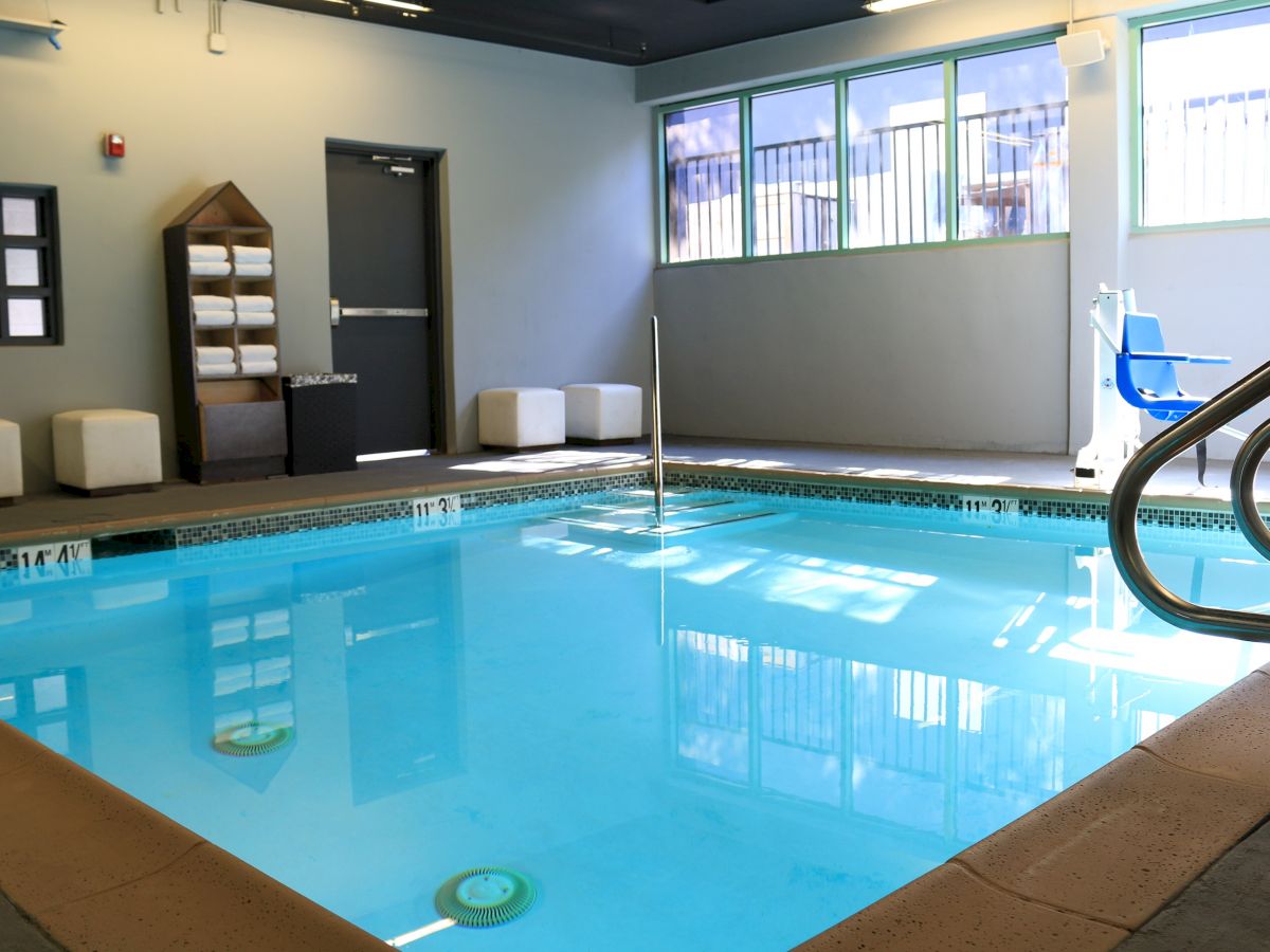 The image shows an indoor swimming pool with a tiled deck, pool lift, towels on shelves, and natural light coming in from windows.