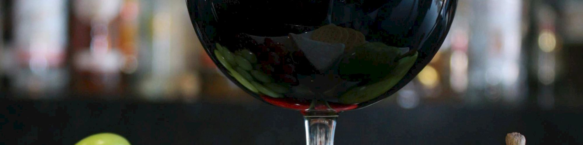 A glass of red wine is in front of a plate with green and red grapes, a pear, an apple, cheese, and crackers, with bottles on a shelf in the background.