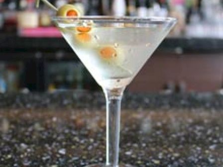 A martini glass filled with a clear cocktail and garnished with green olives on a toothpick is placed on a speckled countertop in a bar setting.