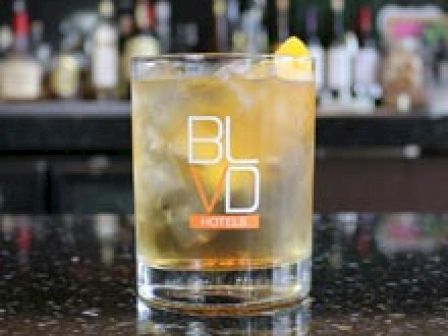 A cocktail glass with a drink and a lemon garnish sits on a bar counter. The glass is labeled 