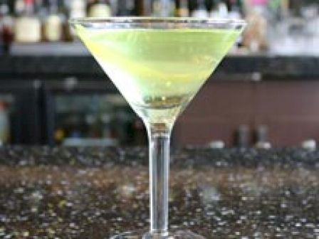 The image shows a martini glass with a yellow cocktail, garnished with a lime slice, placed on a dark speckled countertop in a bar setting.