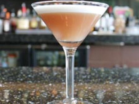 A cocktail in a martini glass on a bar counter with bottles and a blurred background of a bar setting.