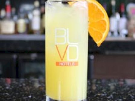 The image shows a tall glass of yellow drink with an orange slice garnish, placed on a bar countertop with 