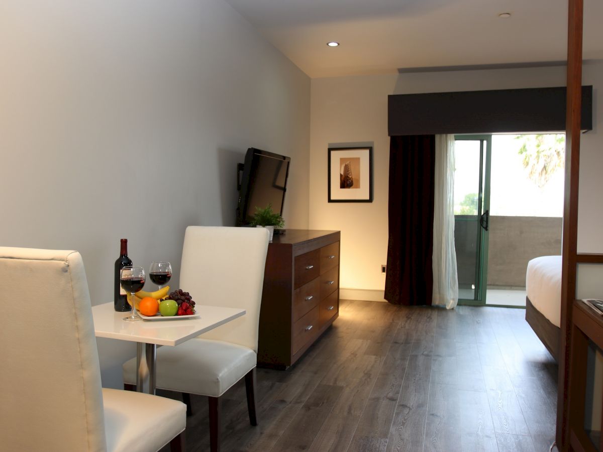 The image shows a modern hotel room with a small dining table and two chairs, a TV, a bed, and a balcony. Wine and fruit are on the table.