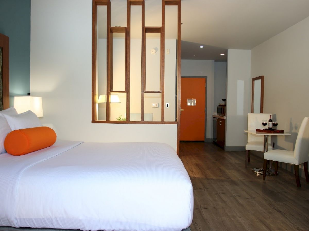 The image shows a modern hotel room with a large bed, an orange pillow, a partition, and a small dining area with a table and chairs on wooden flooring.