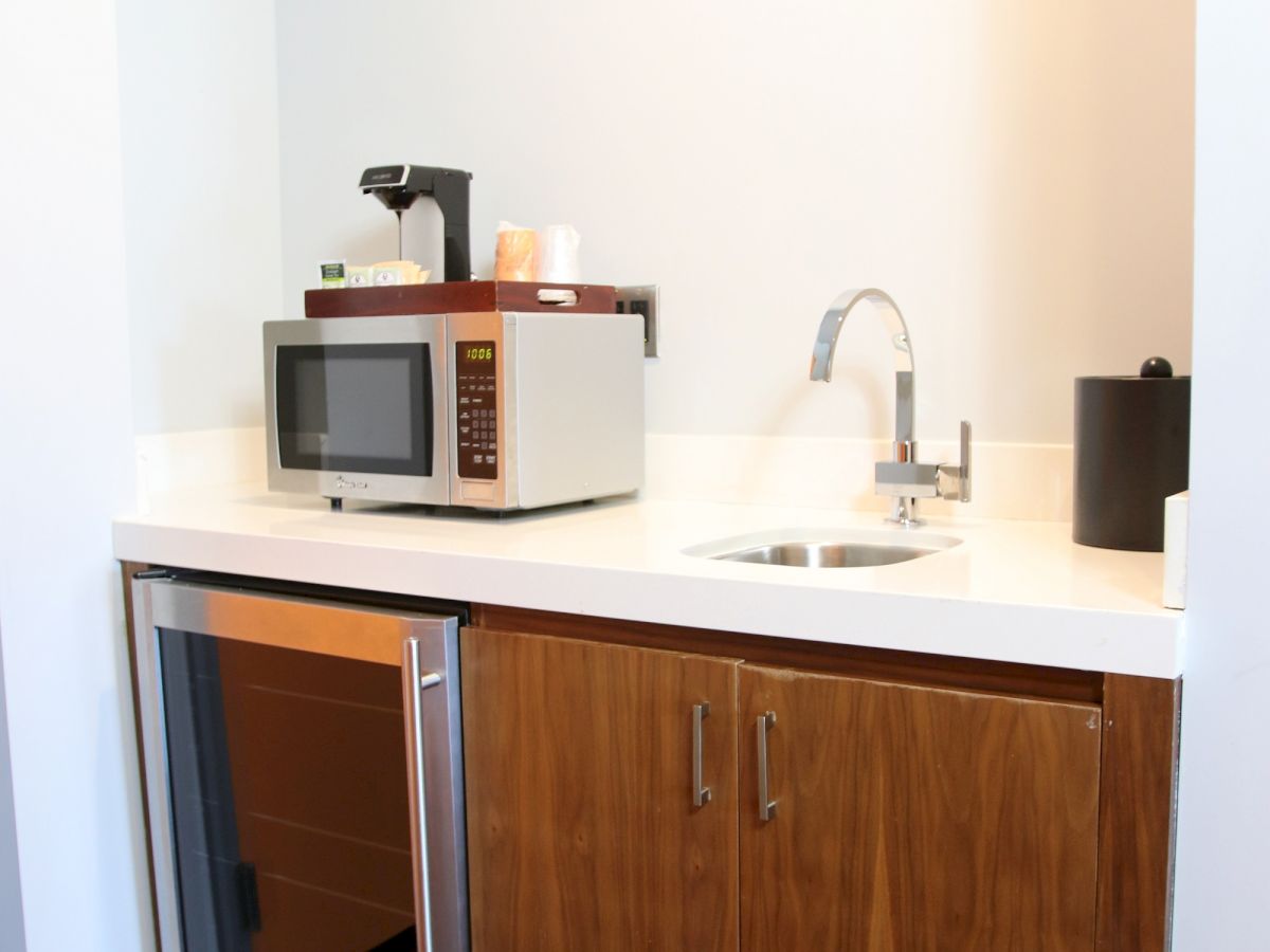 The image shows a small kitchenette with a microwave, a coffee maker, a mini-fridge, a sink, and wooden cabinets.