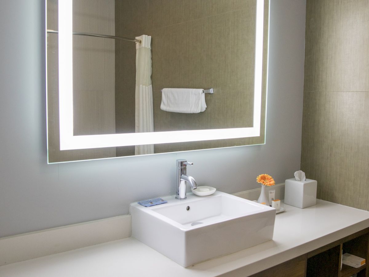 A modern bathroom with a backlit mirror, rectangular sink, faucet, tissue box, and towels on shelves beneath the counter.