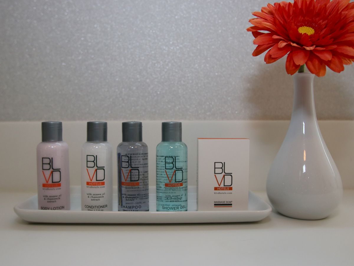 The image shows five BLVD branded toiletry bottles on a tray next to a white vase with an orange flower, on a bathroom counter.