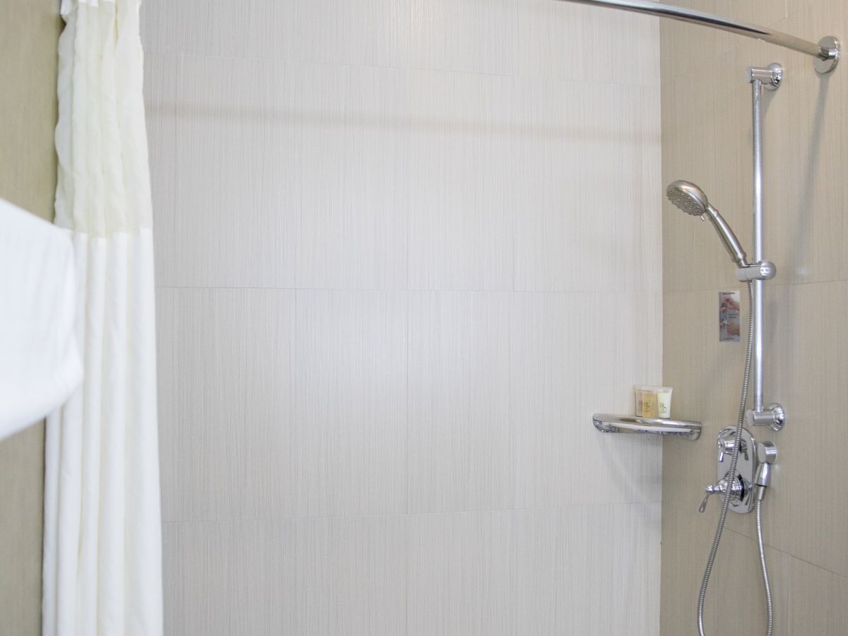 The image shows a clean shower area with a curtain, a wall-mounted showerhead, a handheld shower, and a small shelf holding a candle.