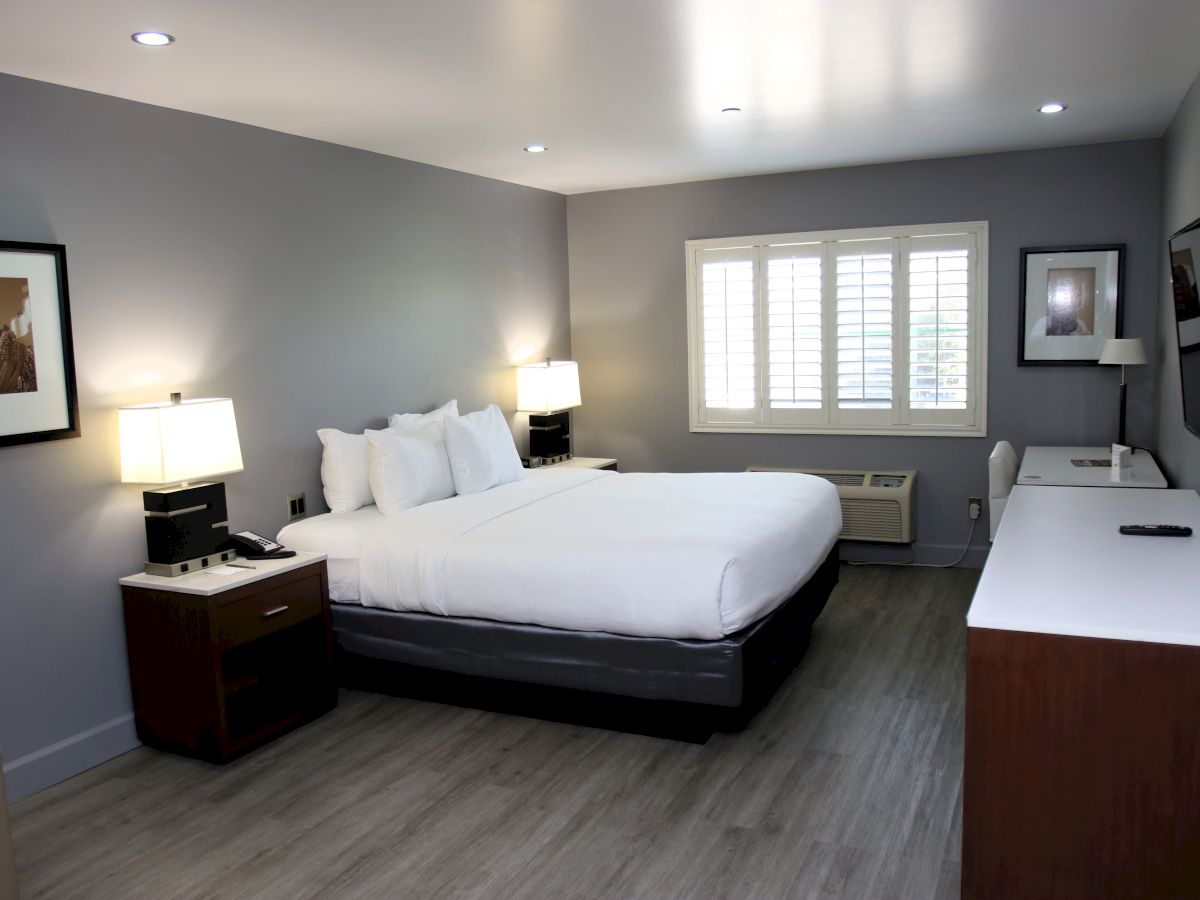 A modern hotel room with a large bed, bedside tables with lamps, a desk, a TV, and wall art, featuring a neutral color scheme.