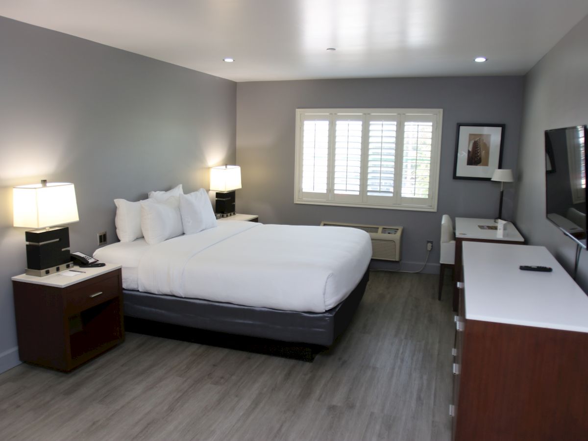 A modern hotel room with a large bed, nightstands, a desk, and a wall-mounted TV. The room has gray walls and wooden floors.