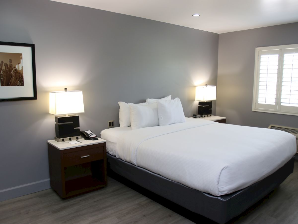 A modern bedroom with a large bed, two nightstands with lamps, a window with shutters, and a framed wall picture.