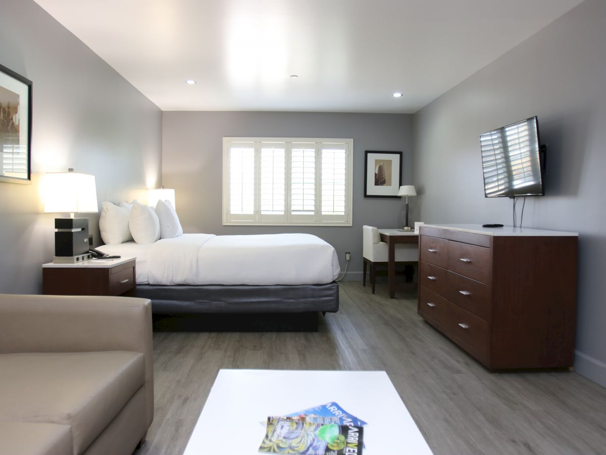 A modern hotel room with a bed, sofa, flat-screen TV, dresser, desk, and window. Bright lighting and simple decor.