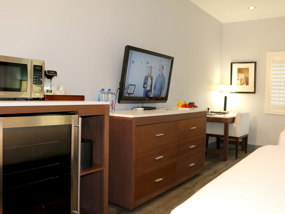 A hotel room features a microwave, mini-fridge, TV on a dresser, desk with a lamp, and a bed on the right.