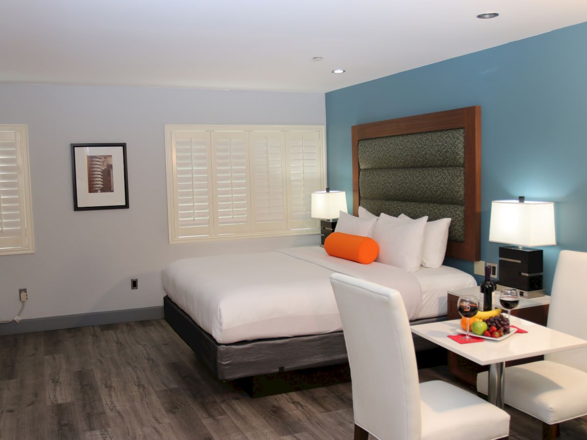 A modern hotel room with a bed, nightstands, lamps, a small dining table with chairs, and a fruit platter, featuring a bright orange cushion.