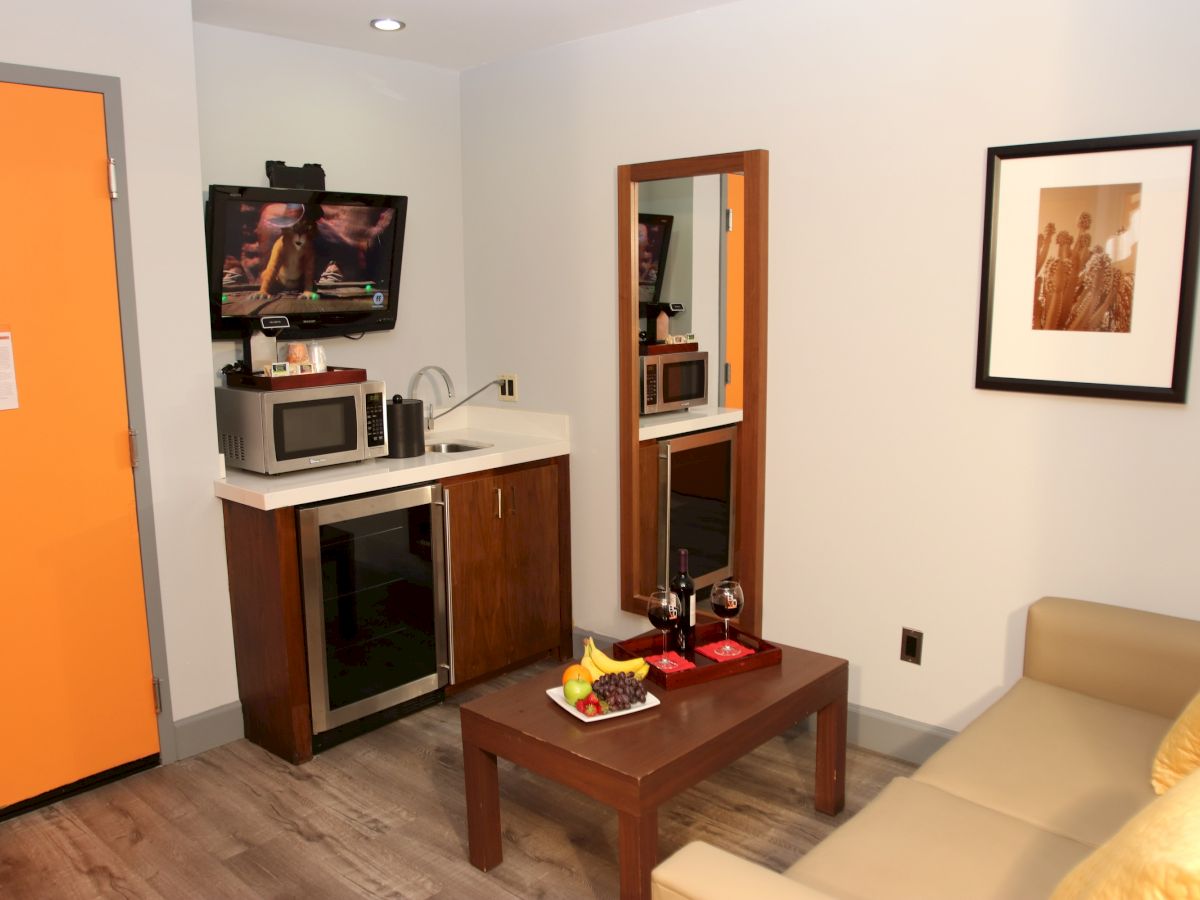 A cozy room with a kitchenette, orange door, TV, microwave, coffee maker, mirror, photo on the wall, beige couch, and coffee table with snacks.