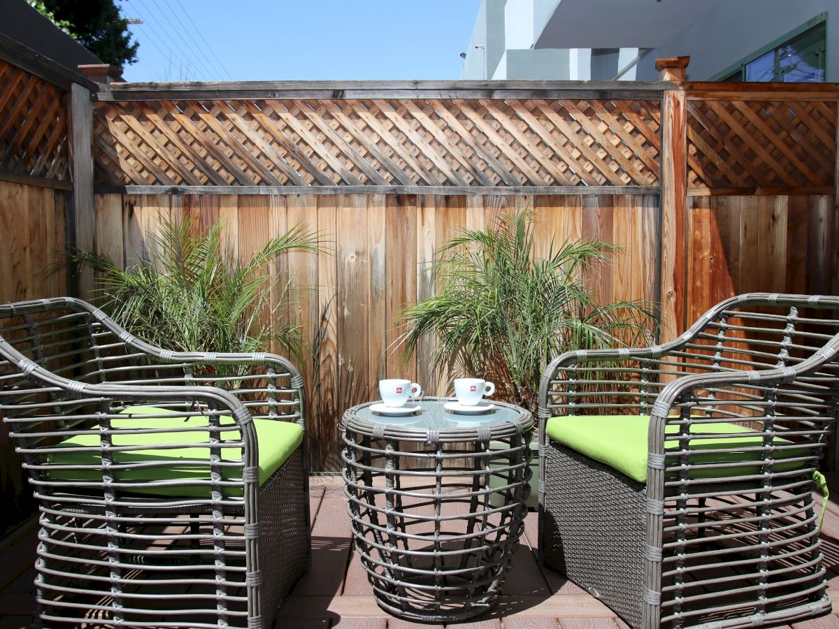 A cozy outdoor patio features two wicker chairs with green cushions, a round matching table with two coffee cups, and potted plants behind the chairs.