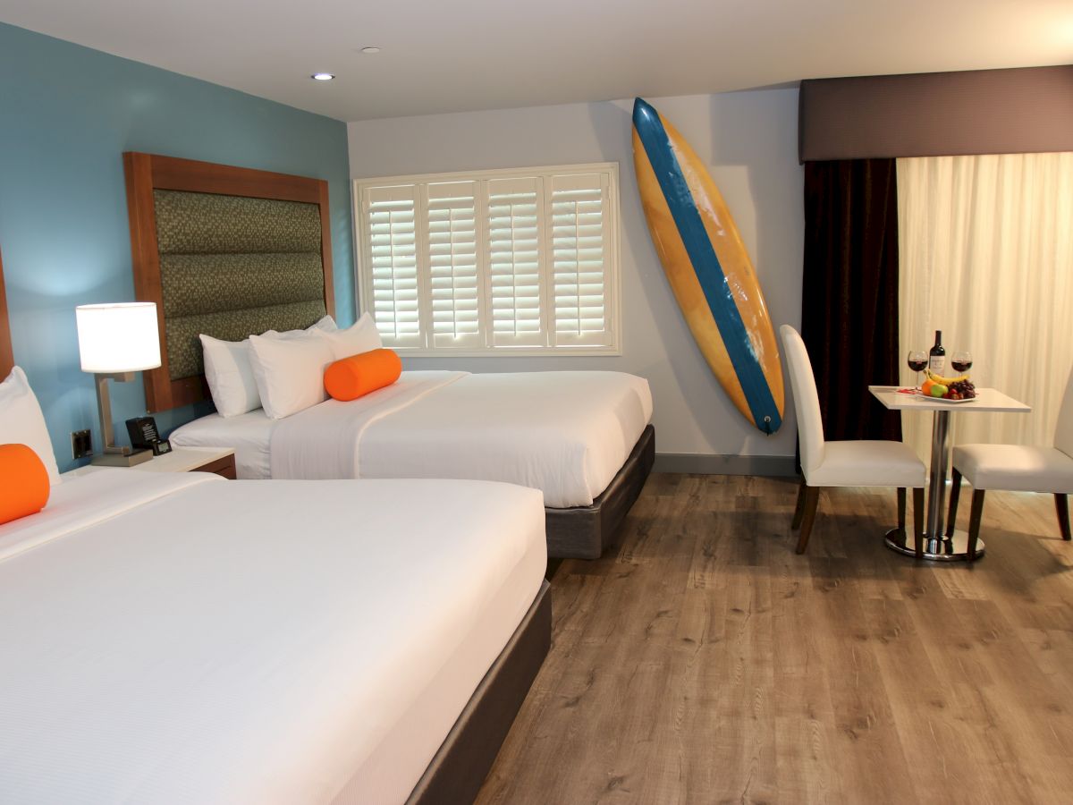 A modern hotel room features two beds, bright decor, a surfboard, a small dining area, and a window with shutters and heavy curtains.