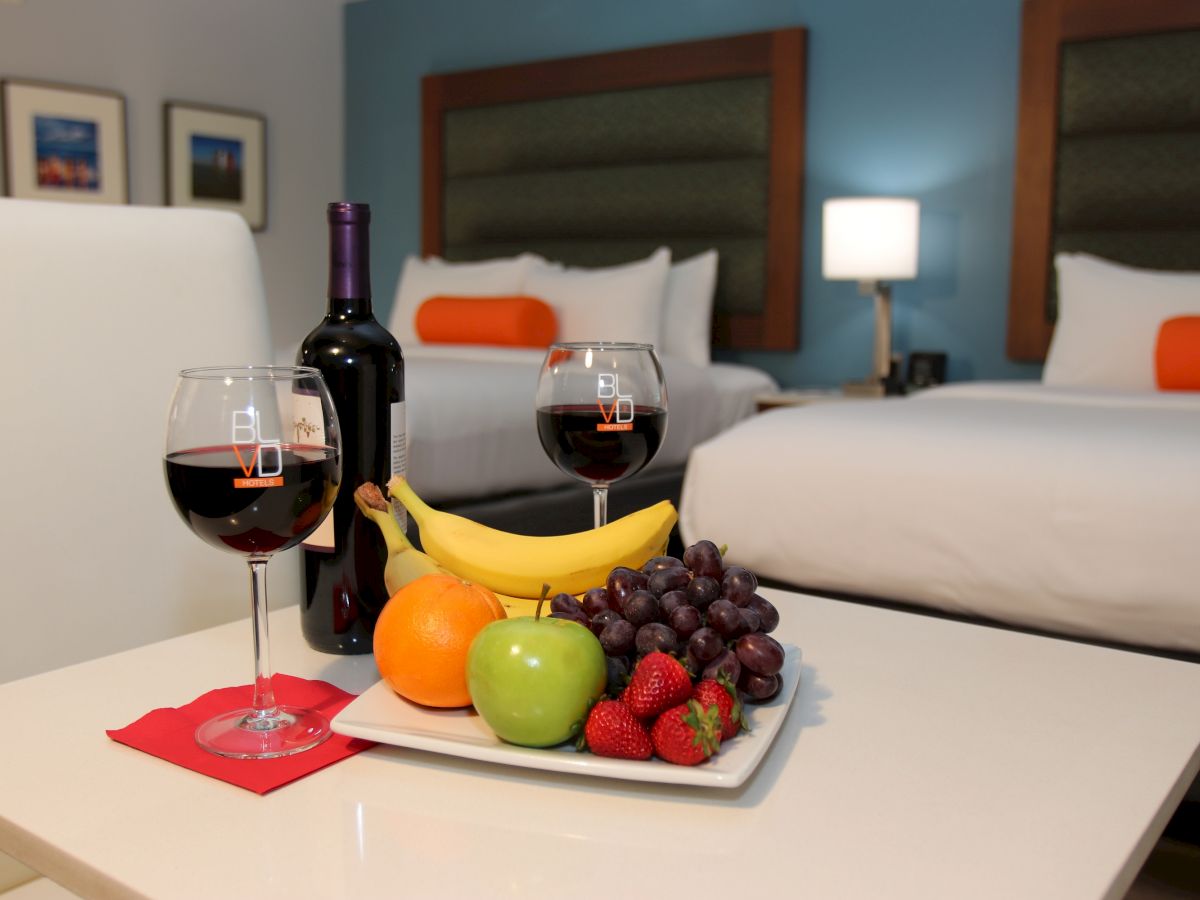 A hotel room features two beds, a lamp, framed art, a bottle of wine, wine glasses, and a fruit plate with bananas, oranges, an apple, grapes, and strawberries.