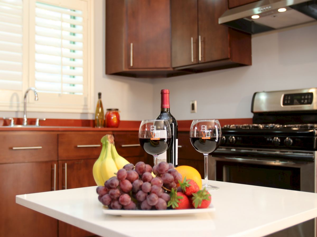 A modern kitchen with wooden cabinets, wine bottle, glasses, plate of fruits including grapes, bananas, strawberries, and an orange on a white counter.