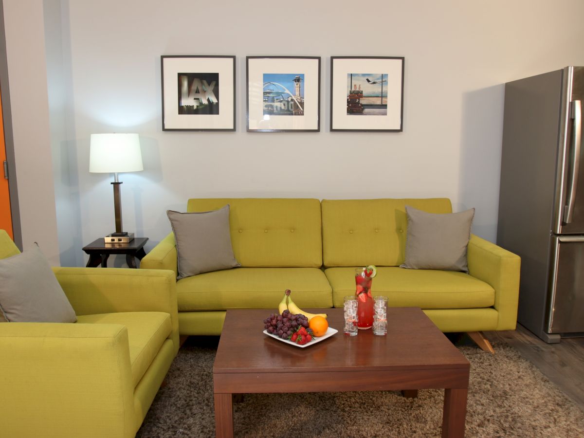 A living room with green sofas, a wooden coffee table, fruit, and drinks. Framed photos on the wall, a lamp, and a fridge are visible.