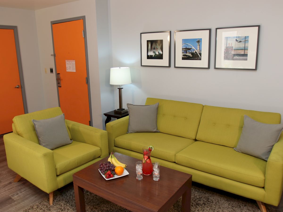 A living room with a green sofa and armchair, a wooden coffee table, and wall art. There's a lamp, fruit, and glasses on the table.