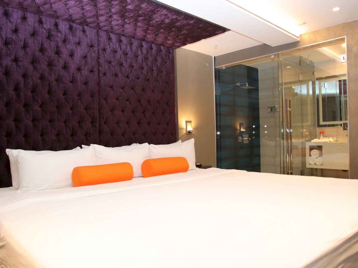 A modern hotel room with a large bed, purple tufted headboard, two orange cushions, and a glass-walled bathroom in the background.