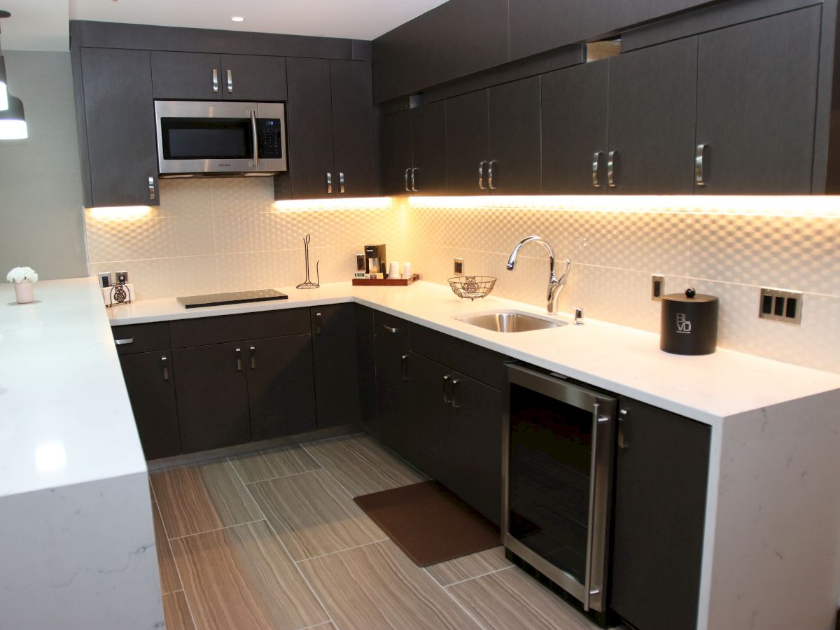 A modern kitchen with dark cabinets, white countertops, a microwave, sink, stove, and under-cabinet lighting.