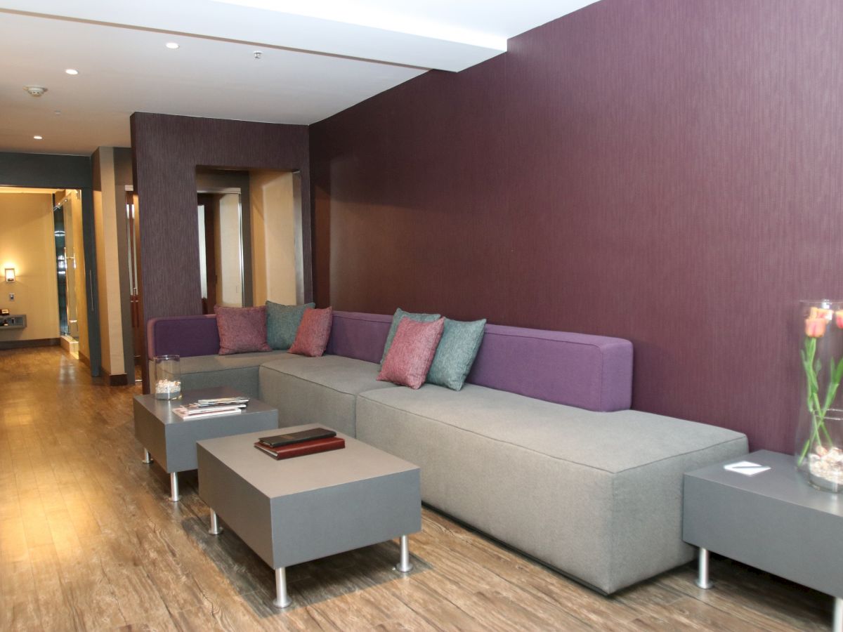 This image shows a modern living room with a long gray and purple sectional sofa, multiple small tables, decorative pillows, and a vase with flowers.