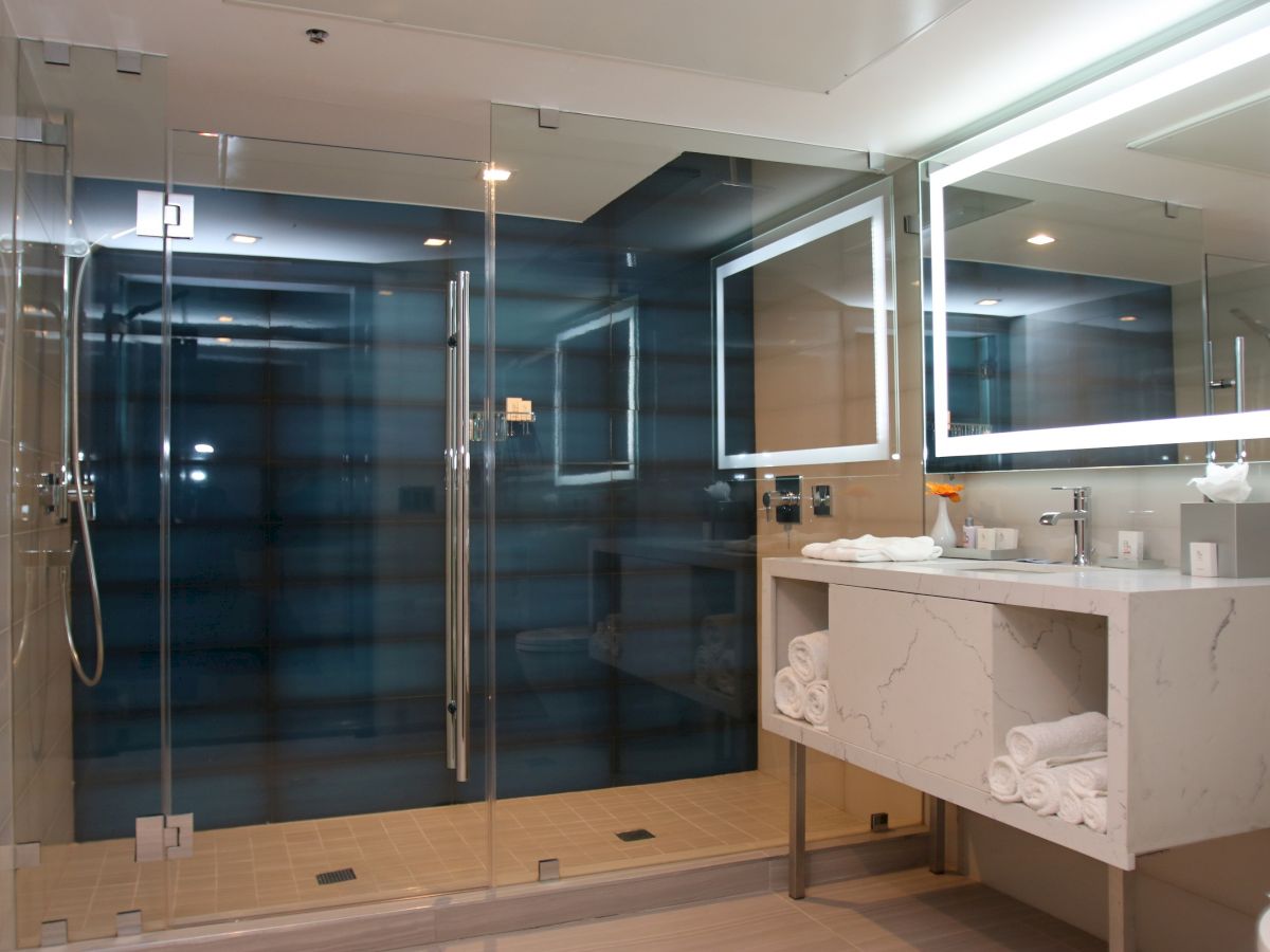 This image shows a modern bathroom with a glass shower enclosure, mirrored vanity with lighting, and neatly organized towels under the sink.