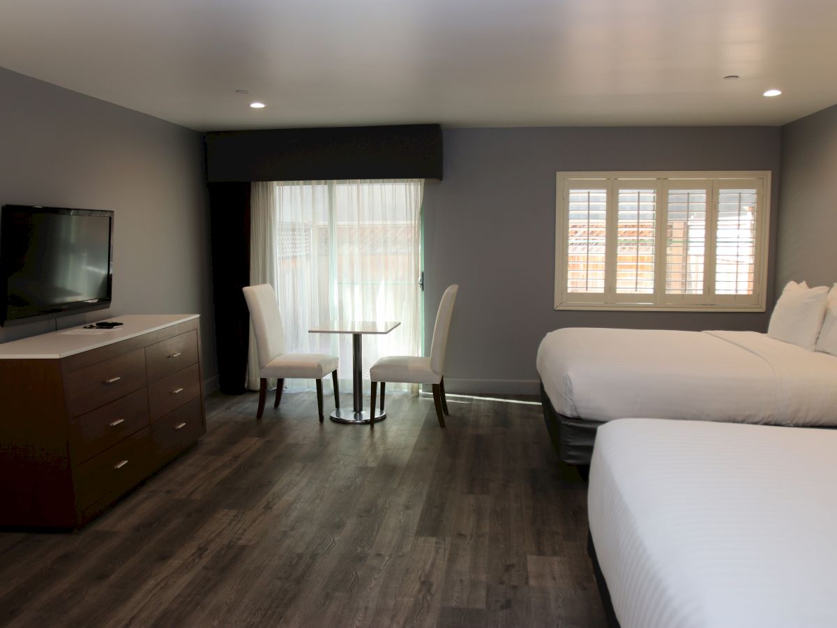 The image shows a hotel room with two beds, a TV on a dresser, a small round table, and two chairs by a window with shutters.