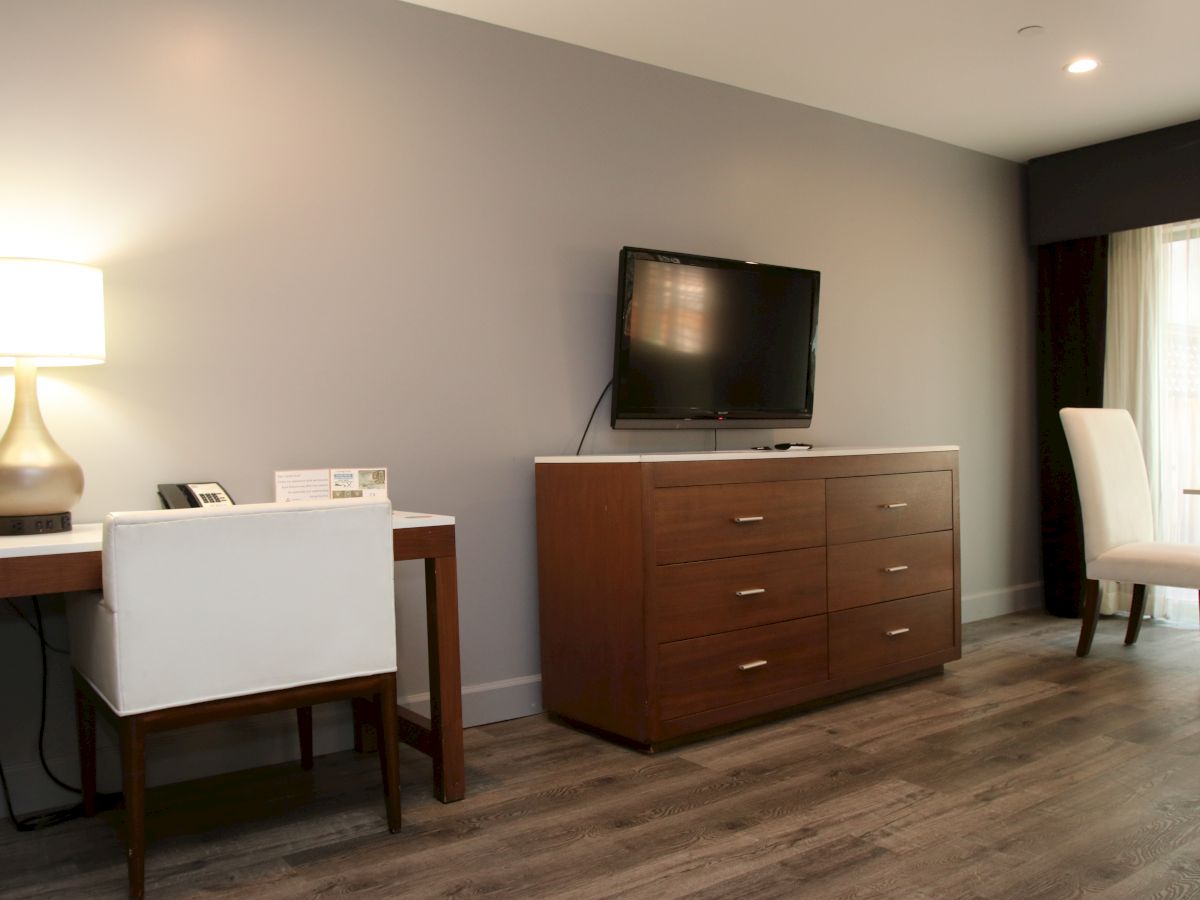 The image shows a modern hotel room with a desk, chair, lamp, dresser, TV mounted on the wall, and a window with curtains.