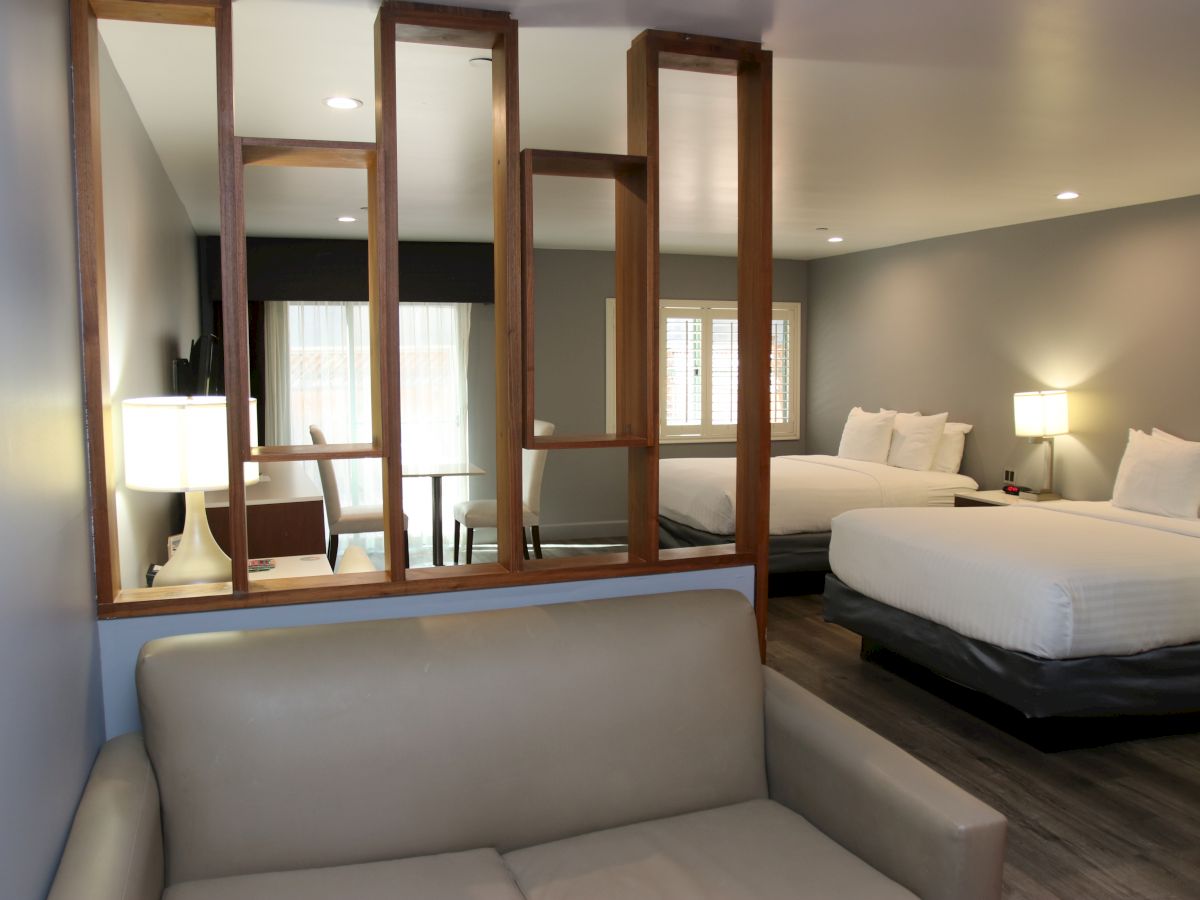 A modern hotel room with a beige sofa, wooden partition, two beds, and soft lighting, creating a cozy and inviting atmosphere.