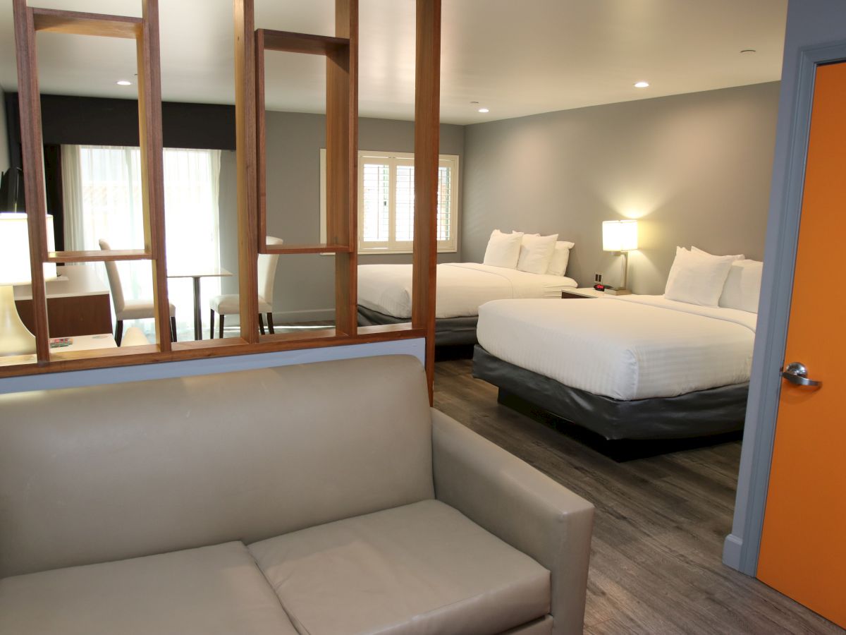 The image shows a modern hotel room with two beds, a gray sofa, a wooden partition, a small table, and an orange door, creating a cozy atmosphere.