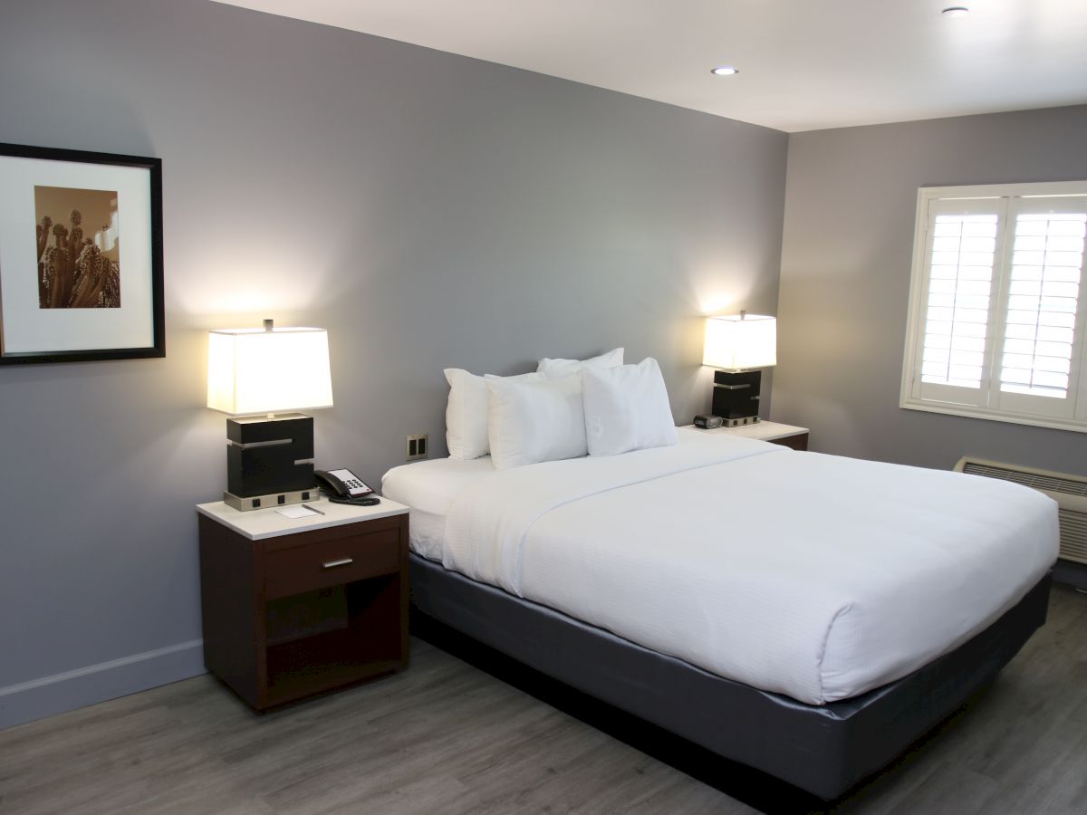 The image shows a modern hotel room with a large bed, nightstands with lamps, art on the wall, and a window with shutters.