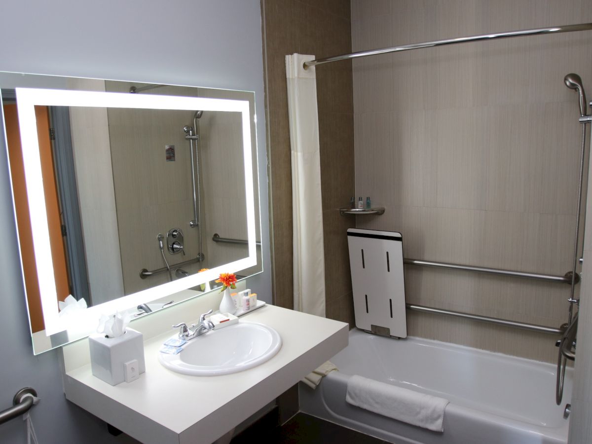 This image shows a modern bathroom with a lit mirror, a sink, a bathtub with a curtain, and a mounted soap dispenser.