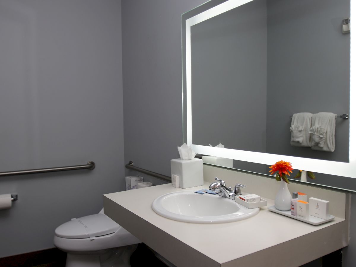 A modern bathroom with a large lit mirror, sink with toiletries, a toilet, and towel rack. There’s a flower vase on the counter.