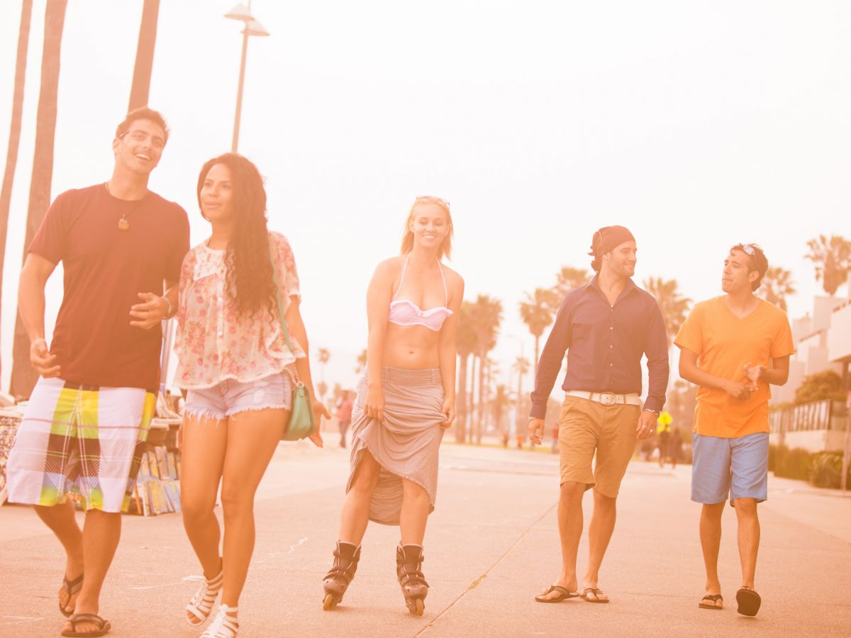 A group of five people walking along a sunny boardwalk lined with palm trees; one person is on rollerblades, and the others seem to be strolling casually.