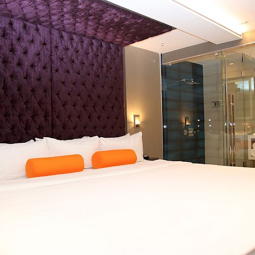 The image shows a modern bedroom with a large bed, purple tufted headboard, two orange pillows, and a glass-walled bathroom in the background.