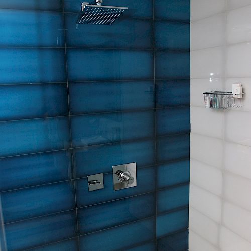 This image shows a modern shower with blue-tiled walls, a rainfall showerhead, and a metal fixture on the right wall holding a soap dish.