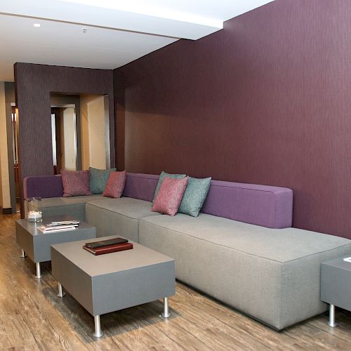 A modern living room with a long gray sofa, purple wall, throw pillows, small tables, and flowers in a vase on the side table.