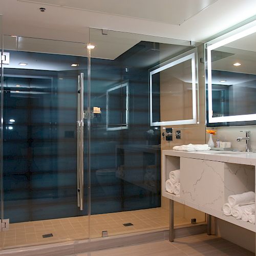 A modern bathroom with a glass-enclosed shower, illuminated mirror, countertop with toiletries, and folded towels on shelves underneath the sink.