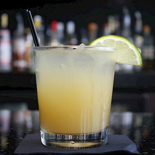 The image shows a cocktail with a lime wedge garnish and a straw, set on a napkin with a blurred bar background.