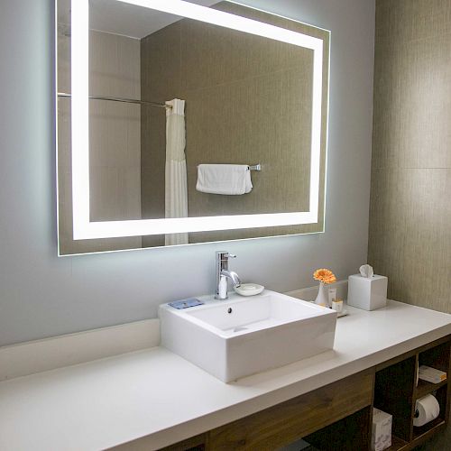 A modern bathroom vanity with a backlit mirror, a rectangular sink, a faucet, a tissue box, and a flower. Towels are stored below the counter.