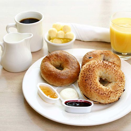 A breakfast setup with bagels, cream cheese, jam, a pitcher of milk, a cup of coffee, orange juice, and a bowl of butter.