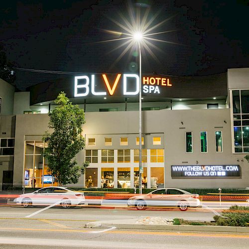 A nighttime street view of the BLVD Hotel & Spa, with cars driving by. The hotel is illuminated, and a sign promotes the website and social media.