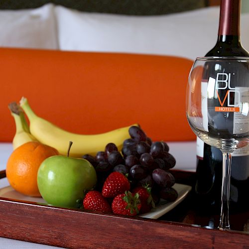 A tray with fruit, a wine bottle, and a glass sits on a bed with an orange pillow in the background.