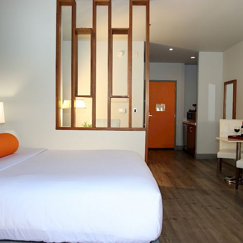 This image shows a modern hotel room with a bed, an orange pillow, a small dining area with chairs, and a kitchenette in the background.