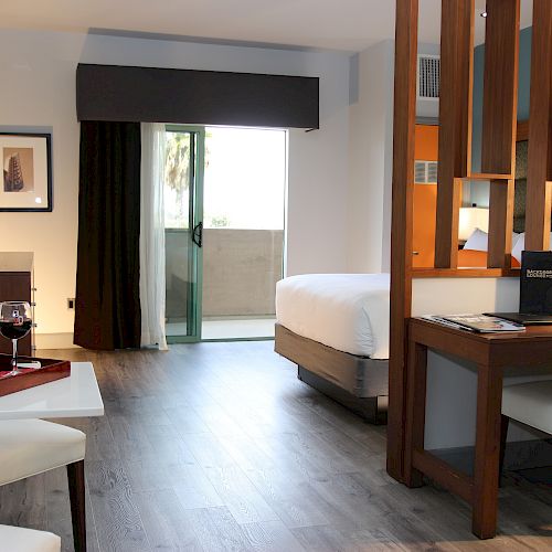 A modern hotel room with a bed, desk, chairs, wine on a table, a balcony window, and stylish room dividers.