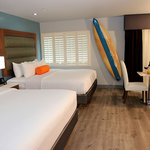 A hotel room features two beds with orange pillows, a surfboard mounted on the wall, a small dining table with two chairs, and a bottle of wine.